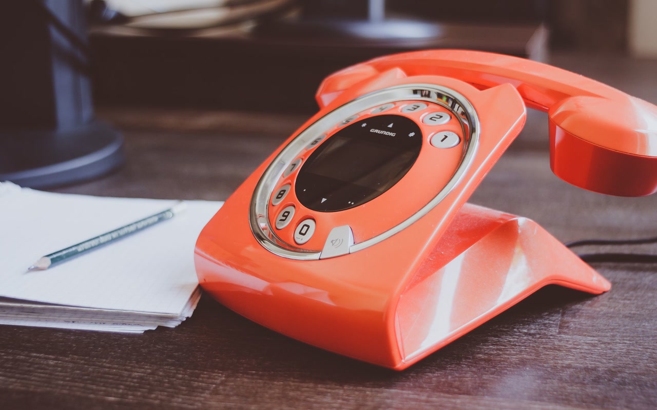 Orange rotary telephone beside a pencil and blank notepad on a wooden desk