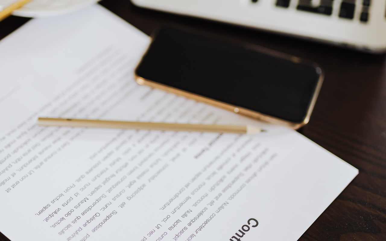 Contract papers, a pen, a smartphone and a laptop arranged on a dark office desk