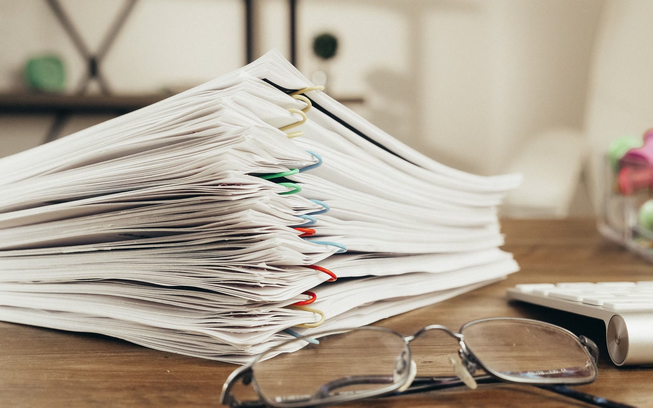 Close-up of stacked paper documents held together with coloured clips, with a pair of reading glasses resting beside them on a desk.