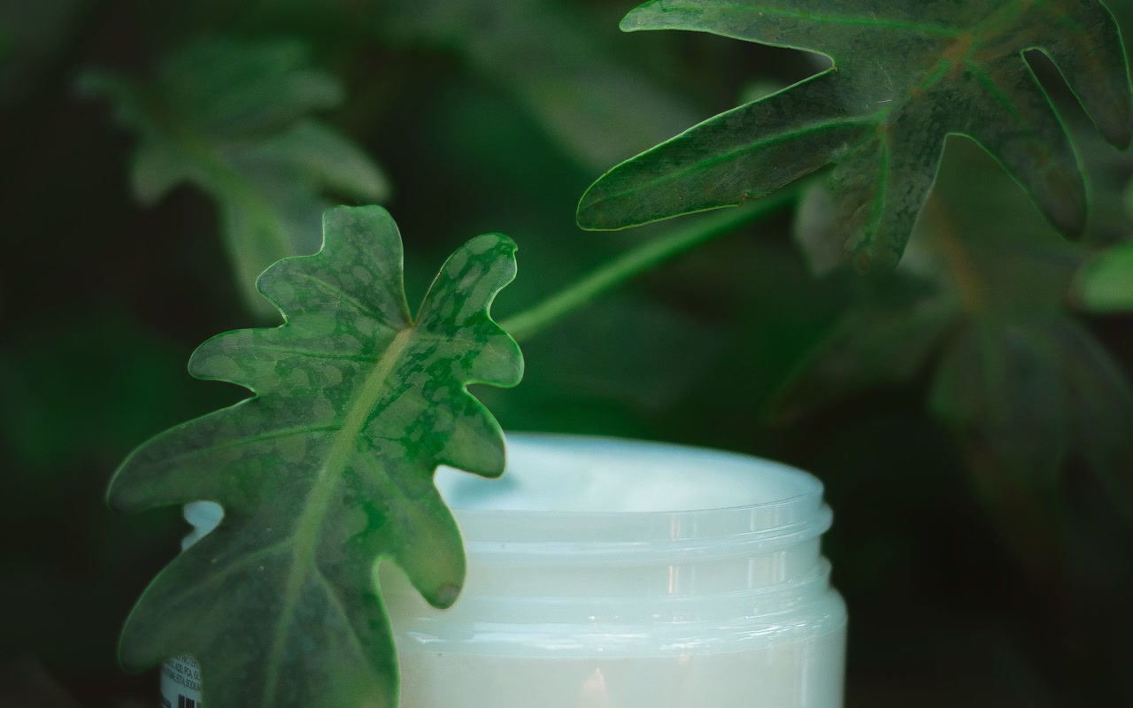 Unlabelled white cosmetic cream jar beside green leaves on a neutral surface