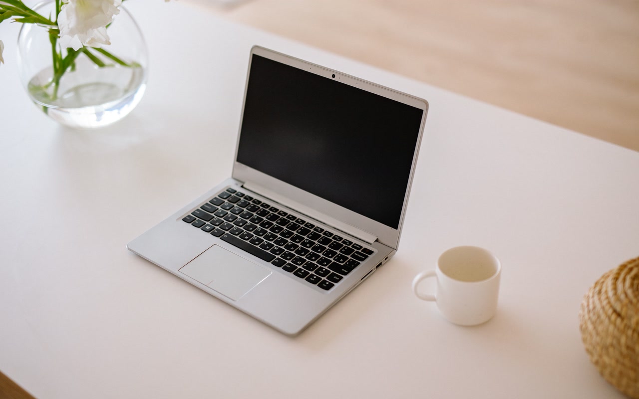 An open silver laptop with a blank screen, a white mug and a clear glass vase of white flowers arranged on a bright white table, viewed from above.
