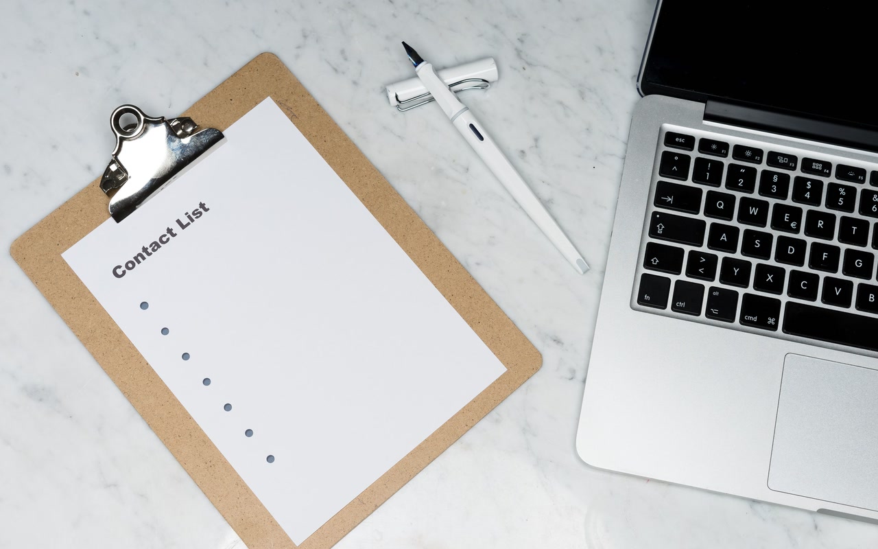 A blank contact-list sheet on a clipboard beside a pen and an open laptop on a marble desk.