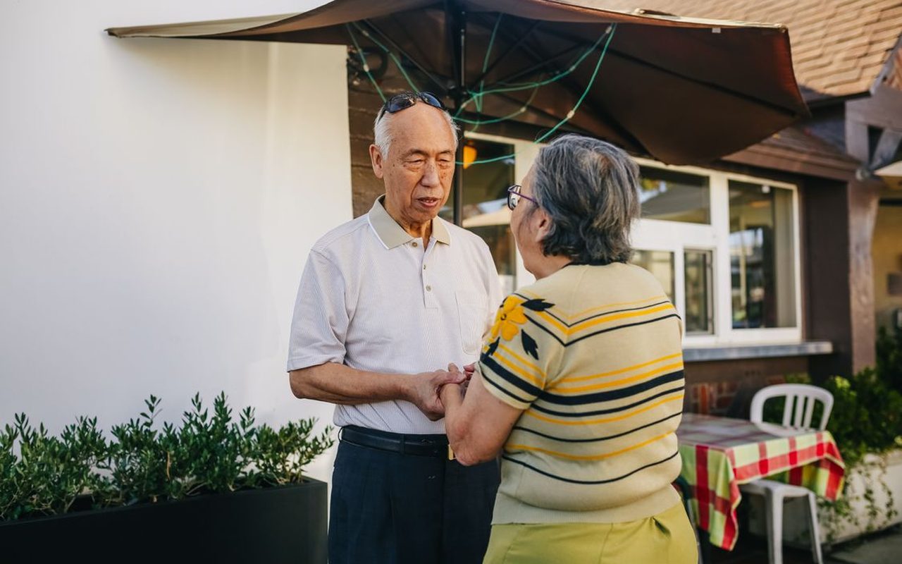 An older couple standing outdoors talking, holding hands under a cafe umbrella