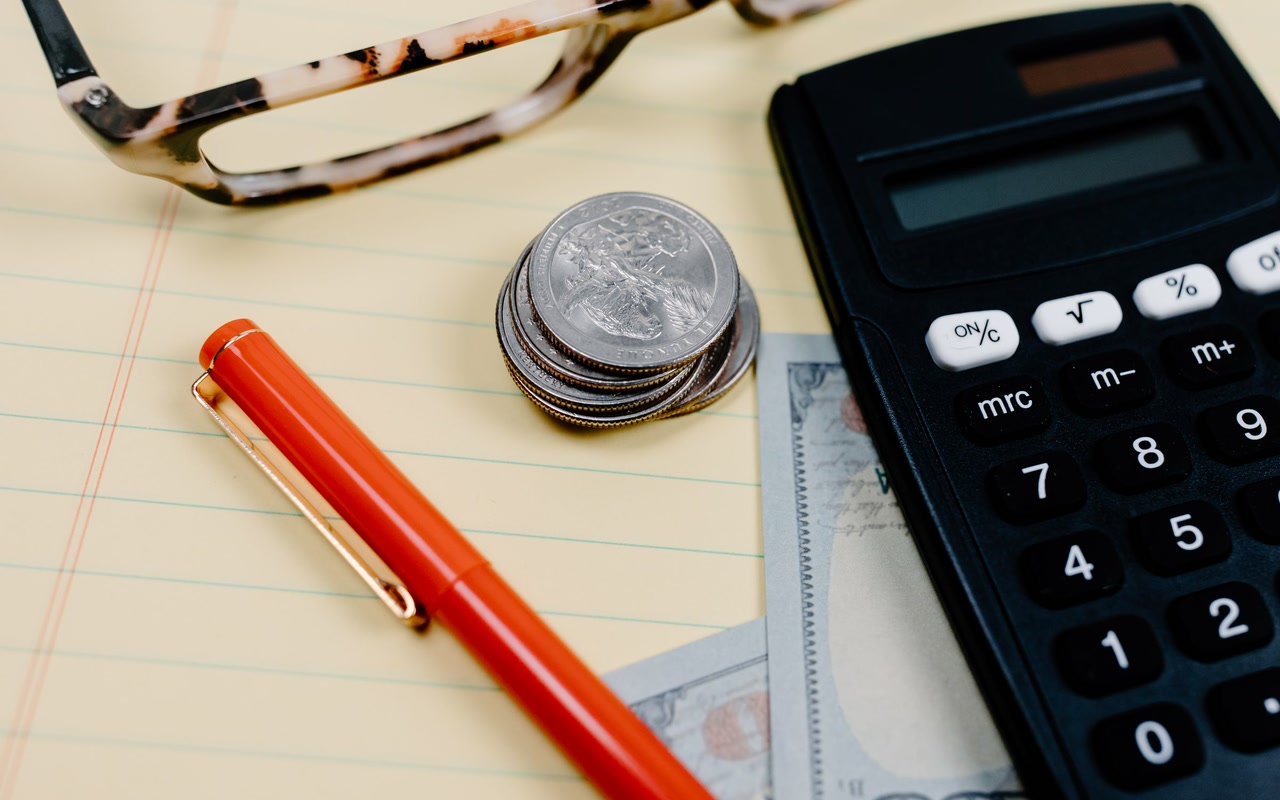 Calculator, red pen, loose coins and glasses arranged on a blank lined notepad