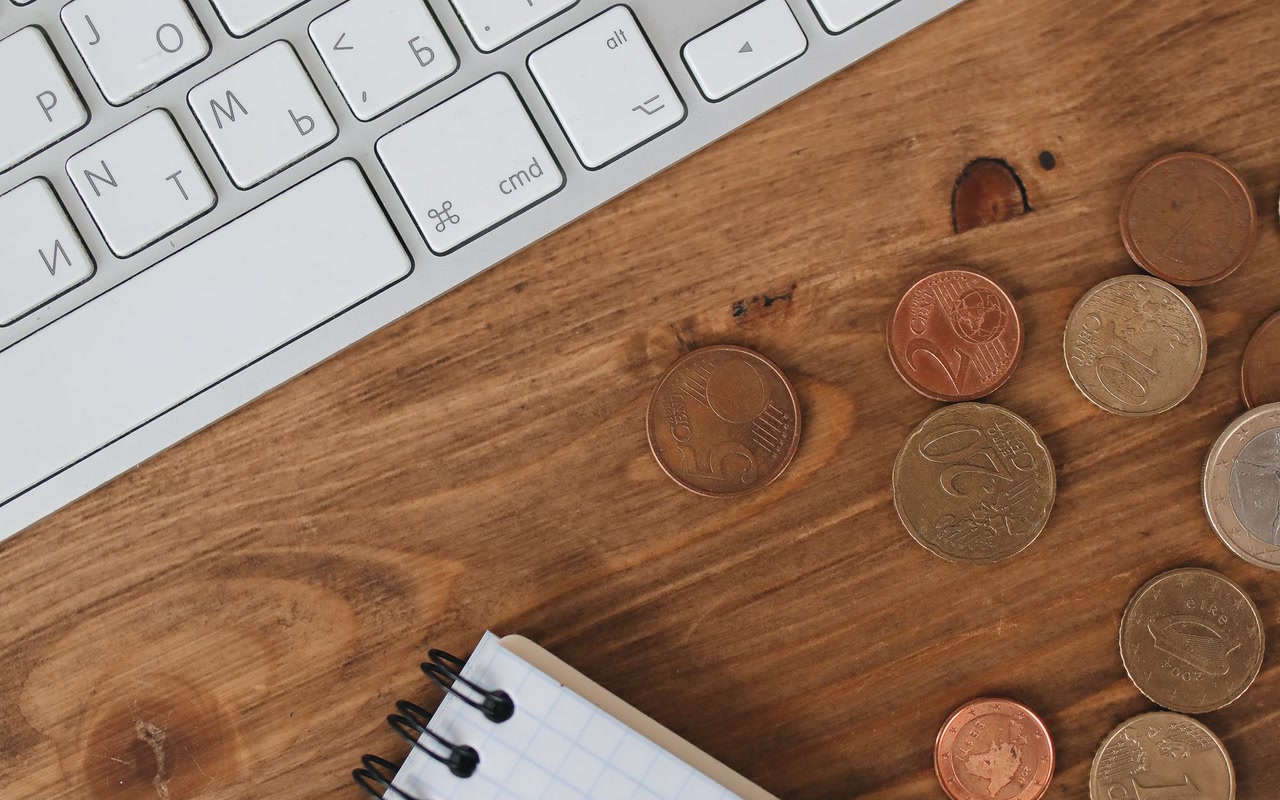 Keyboard, coins and a blank notebook arranged on a wooden work desk