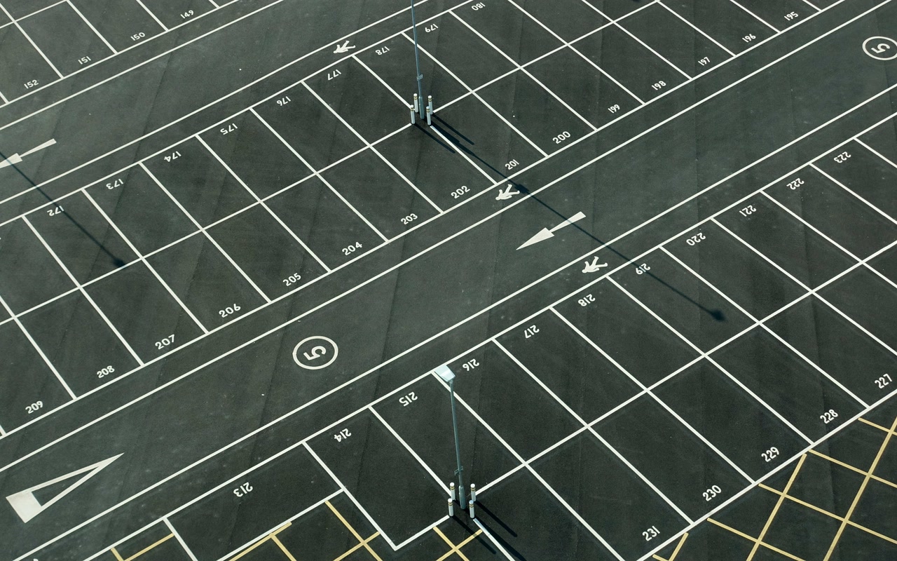Aerial view of an empty asphalt car park with white painted bay markings and numbered spaces, with light poles casting long shadows.